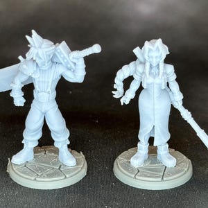 May include: Two light blue figurines of video game characters, one holding a large sword and the other holding a staff. Both figures are standing on gray circular bases with a stone-like texture. The figures are detailed and appear to be 3D-printed.