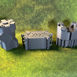May include: Three gray, 3D-printed miniature structures are arranged on a green surface. The structures include a tall, faceted tower, a rectangular building with intricate details, and a building with a curved design. The objects appear to be game pieces.
