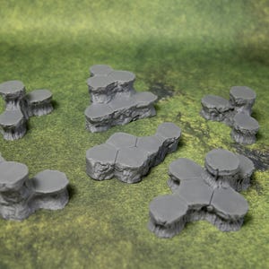 May include: A collection of gray, multi-tiered rock formations, likely for tabletop gaming or model scenery. The pieces vary in size and shape, with a textured surface, and are arranged on a green, textured surface.