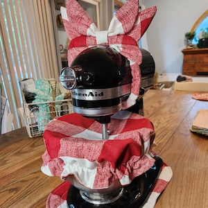 May include: A black KitchenAid Classic stand mixer adorned with a red and white gingham fabric cover, headband, and bowl cover. The accessories create a cohesive, decorative look. The mixer sits on a wooden table.