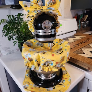 May include: A black KitchenAid stand mixer with a yellow bee-themed cover and bow. The cover and bow feature a repeating pattern of bees, flowers, and clouds on a yellow background. The mixer is on a white countertop.