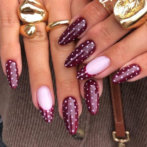 Peut inclure: Ongles en amande bordeaux avec des pois blancs. Certains ongles ont une base rose clair. Des bagues dorées ornent les doigts. Les ongles sont d'un rouge profond avec une finition brillante, et les pois sont espacés uniformément.