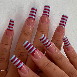 Peut inclure: Longs ongles carrés avec une base transparente et un design de french manucure. Chaque ongle est orné de rayures horizontales alternées bordeaux et bleu clair, créant un motif audacieux. Les ongles sont bien manucurés et polis.