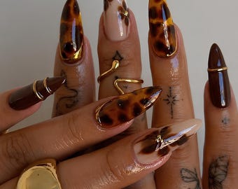 Puede incluir: Uñas postizas en forma de almendra con una mezcla de patrones brillantes de carey, esmalte marrón oscuro sólido y bases nude con diseños florales marrones pintados a mano. El conjunto incluye detalles metálicos dorados y tachuelas doradas.