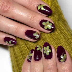 Può includere: Primo piano di unghie curate con smalto bordeaux scuro. Ogni unghia presenta un delicato motivo floreale verde chiaro. Le unghie sono di forma ovale e hanno una finitura lucida, su uno sfondo color senape.