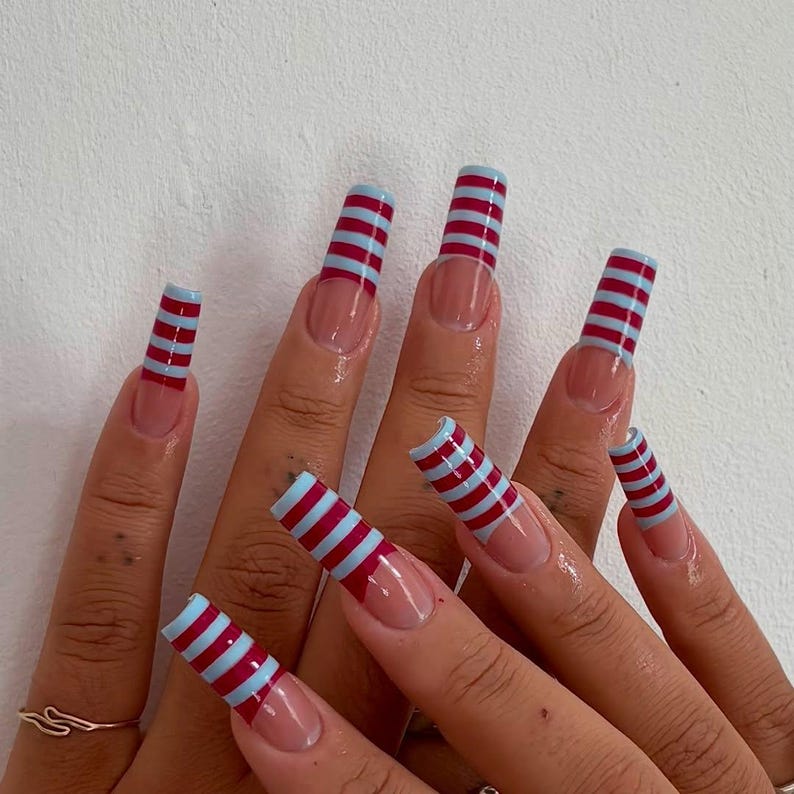 May include: Long, square-shaped nails with a clear base and a French tip design. Each nail is adorned with alternating horizontal stripes of burgundy and light blue, creating a bold and eye-catching pattern. The nails are well-manicured and polished.