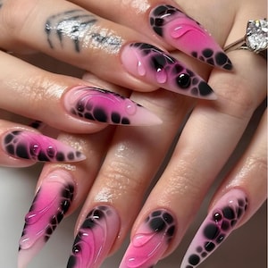Può includere: Unghie lunghe e appuntite con un design sfumato. Le unghie passano da un rosa pallido alla base a un rosa vibrante al centro, con motivi neri a forma di bolla e accenti trasparenti a forma di goccia. È visibile un anello con una grande pietra trasparente.