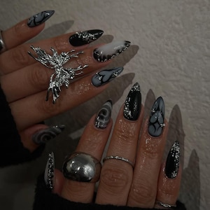 Puede incluir: Uñas largas y puntiagudas con una combinación de colores oscuros. Las uñas presentan diseños en negro, gris y blanco, con detalles plateados y un anillo de mariposa plateada. Las uñas tienen un acabado brillante.