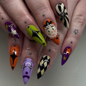 Puede incluir: Una manicura con uñas largas y puntiagudas, decoradas con diversos diseños. Las uñas presentan una cara de payaso, estrellas y patrones geométricos en colores como naranja, morado, verde y negro. Algunas uñas tienen un diseño a rayas blanco y negro.