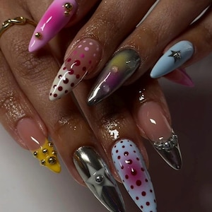 Puede incluir: Uñas largas en forma de almendra con diseños variados que incluyen degradado rosa con tachuelas doradas, blanco con lunares rojos, plata metálica con estrellas, azul claro con un dije de estrella y amarillo con tachuelas doradas.