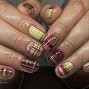 Op de afbeelding: Close-up van gemanicuurde nagels met een ruitpatroon. De nagels hebben een mix van kleuren, waaronder geel, bordeauxrood en bruin, gerangschikt in een klassiek ruitpatroon. De nagels zijn ovaal van vorm en hebben een glanzende afwerking.