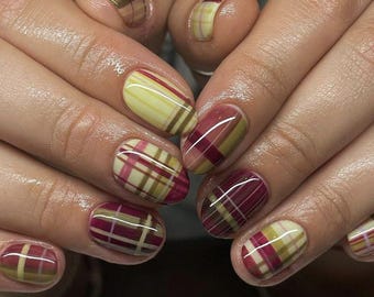 Puede incluir: Primer plano de uñas manicuradas con un diseño de cuadros. Las uñas presentan una mezcla de colores, incluyendo amarillo, burdeos y marrón, dispuestos en un patrón de cuadros clásico. Las uñas son ovaladas y tienen un acabado brillante.