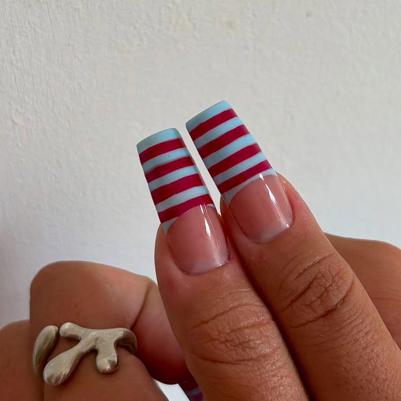May include: Close-up of two long, square-shaped fingernails with a light pink base and a French tip. The nails are painted with alternating horizontal stripes of light blue and red. A silver ring with an abstract design is visible.