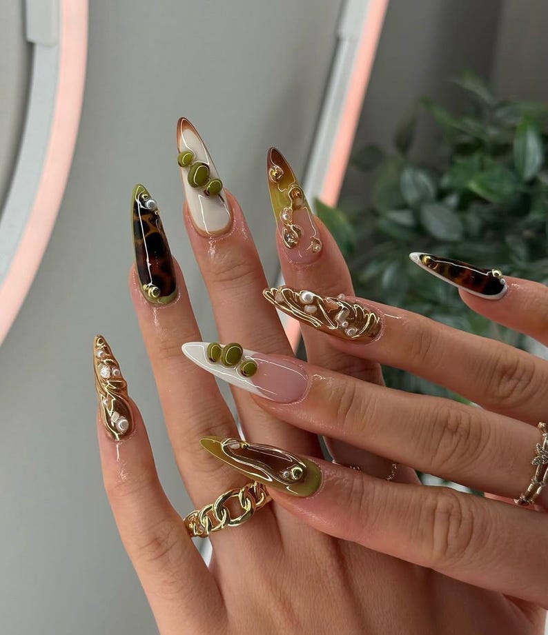 Puede incluir: Primer plano de una mano con u&ntilde;as acr&iacute;licas largas en forma de almendra. Las u&ntilde;as presentan una variedad de dise&ntilde;os, incluyendo patrones de carey, puntas blancas y acentos verde oliva. Algunas u&ntilde;as est&aacute;n adornadas con adornos dorados y perlas.