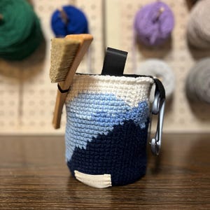 Puede incluir: Un saco de tiza de ganchillo en tonos azules y blancos, con un lazo negro y un mosquetón plateado. Un cepillo de madera está insertado en la bolsa. Diseñado para escalada.