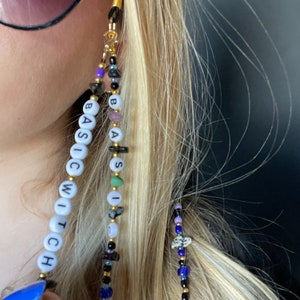 May include: A pair of glasses chain featuring the words "BASIC WITCH" in white beads, with black, blue, purple, and green beads interspersed. The chain is attached to a gold-coloured clasp.