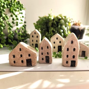 Puede incluir: Un conjunto de pequeñas casas de cerámica en tonos beige y marrón, cada una con detalles de puertas y ventanas negras. Las casas están dispuestas sobre una superficie blanca, con plantas verdes en el fondo.