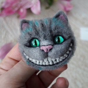 Könnte beinhalten: Eine graue Filz-Brosche mit einem breiten Grinsen, grünen Augen und einer rosa Nase, die den Cheshire Cat darstellt.