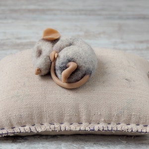 Puede incluir: Una pequeña figurita de ratón de fieltro gris y marrón acurrucada y durmiendo sobre una almohada beige con un borde cosido morado.