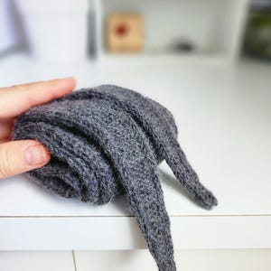 Puede incluir: Una diadema de punto gris con orejas largas y caídas. La diadema está doblada y colocada sobre una superficie blanca.