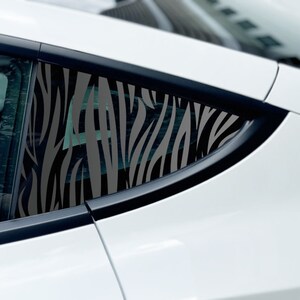 May include: A dark gray zebra stripe pattern decal applied to the triangular rear side window of a white car. The vinyl graphic features elongated, irregular vertical lines that follow the shape of the glass panel on the vehicle exterior.