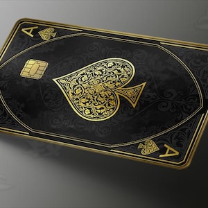 May include: A rectangular payment card featuring a black background with intricate gold filigree patterns. The center displays a large gold spade symbol, while the corners show the letter A and smaller spade icons. A gold EMV chip is visible.