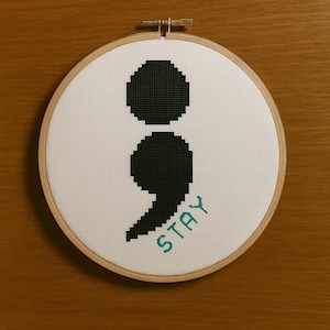 Può includere: Un telaio da ricamo a punto croce con un disegno a punto e virgola nero su tessuto bianco. La parola "STAY" è ricamata in turchese sotto il punto e virgola. Il telaio in legno è posto su uno sfondo di legno marrone.