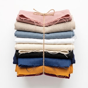 May include: A stack of folded fabric napkins in various colors including dusty rose, beige, slate blue, white, cream, black, navy, mustard yellow, and deep burgundy, all tied together with a thin brown twine string in a simple bow.