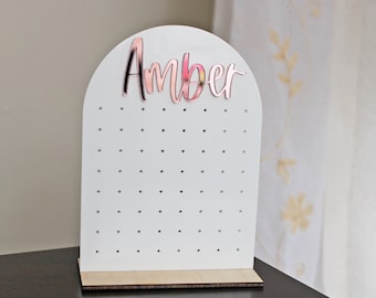 May include: White earring holder with a rounded top and a wooden base. The holder has a grid of holes for storing earrings. The name "Amber" is written in rose gold lettering on the top of the holder.