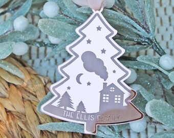 May include: A rose gold Christmas tree ornament with a white silhouette of a house, trees, a crescent moon, and stars. The ornament has the text "The Ellis Family" at the bottom.