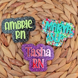 May include: Three colorful glitter name tags with the names "Ambrie RN", "Marya SLP", and "Tasha RN" on a woven wicker background.