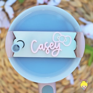 Pode incluir: Um copo reutilizável azul claro com uma tampa rosa e um canudo. A tampa tem um rótulo branco e verde com o nome "Casey" escrito em itálico rosa e um laço rosa.