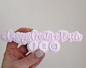 May include: A custom social media sign featuring the text @CaseysCreativeTouch in white raised lettering on a light pink background. Below the text are three circular icons representing Facebook, Etsy, and Instagram in white and pink.