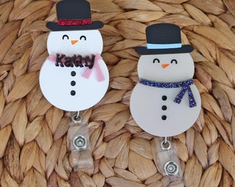 May include: Two snowman-shaped badge reels. Each snowman has a black top hat, a carrot nose, and a scarf. One has the name "Kathy" on a pink scarf. The other has a blue scarf. Both have black button eyes and a black mouth.