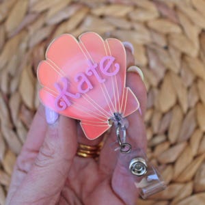 Puede incluir: Un portatarjetas en forma de concha marina en tonos coral y rosa, con el nombre "Kate" en escritura rosa. El portatarjetas tiene un clip de plástico transparente y un cordón retráctil.