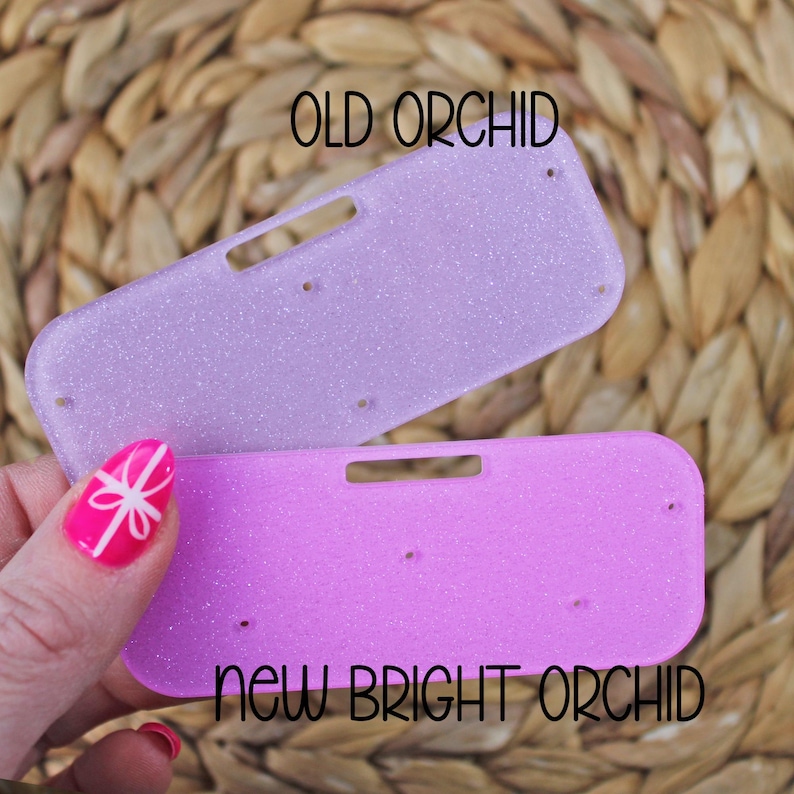 May include: Two glittery, rectangular accessories in shades of purple and pink. The top one is a lighter lavender color, labeled "OLD ORCHID," while the bottom is a brighter pink, labeled "NEW BRIGHT ORCHID." Each has a rectangular cutout and several small holes.