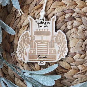 May include: A wooden ornament in the shape of a semi-truck with angel wings. The ornament has the text "Trucking in Heaven" at the top, the name "David" in the middle, and the year "2024" at the bottom. It hangs from a white string.