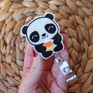 May include: A cute panda bear badge reel with a black and white embroidered design. The panda is holding a candy corn. The badge reel has a clear plastic retractable cord and a metal clip.
