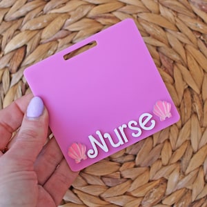 May include: A pink nurse badge holder with a slot for a lanyard. The word "Nurse" is written in white script, with two pink seashell accents. The badge holder is held by a hand with light purple nail polish.