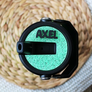May include: Black water bottle with a green speckled design and the name "AXEL" in black letters. The bottle has a black handle and a black spout. The background is a woven, light brown surface.