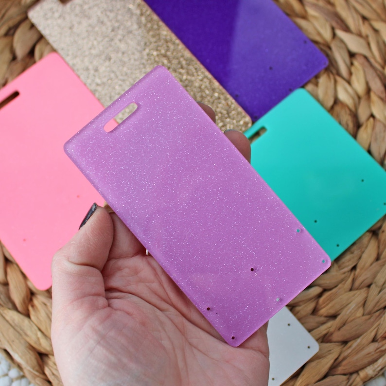 May include: A hand holding a rectangular, lavender-colored earring display card with glitter. Other earring display cards in pink, gold glitter, purple, teal, and white are visible in the background. Each card has a hole at the top and several holes for earrings.