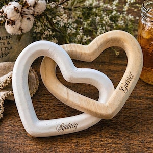May include: Two interlocking heart-shaped wooden sculptures. One heart is painted white with the name Sydney engraved in script, and the other is a natural light wood finish with the name Garret engraved in script on the side of the heart.