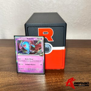 May include: A Team Rocket's Wobbuffet Pokémon card with a blue creature illustration, next to a black and red storage box with a large white "R" logo. The card has text that reads "Rocket Mirror" and "Headbutt Bounce".