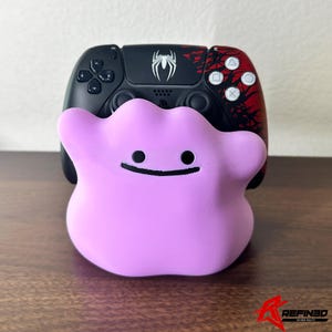 May include: A pink Ditto figure with a black smiling face is in front of a black and red PlayStation 5 controller. The controller has a white spider logo and red accents.