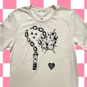 Puede incluir: Una camiseta de color crema con un gráfico negro de un corazón con púas con una cadena y una estrella en la cadena. El texto "APRIL WTC" está en un corazón negro debajo del gráfico.