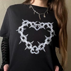 Puede incluir: Camiseta negra con un diseño blanco en forma de corazón con eslabones de cadena y púas. Se lleva un collar plateado con abalorios. Las mangas están superpuestas a una camiseta de manga larga a cuadros en blanco y negro.