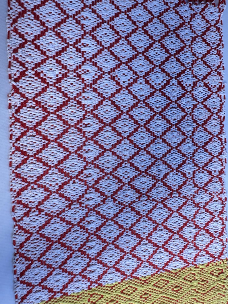 Puede incluir: Una alfombra tejida roja y blanca con un patr&oacute;n de diamantes. El borde inferior de la alfombra es amarillo.