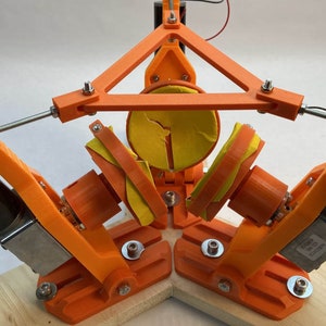 May include: A 3D printed orange and yellow robotic arm with three motors and a circular platform. The platform is attached to the arms by rods and is partially covered in yellow foam.