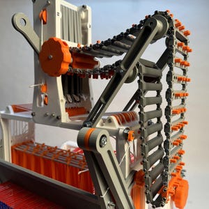May include: A close-up of a 3D printed loom with a chain mechanism. The loom is made of white plastic with orange accents. The chain is made of gray plastic and has orange teeth. The loom is weaving a red and blue fabric.