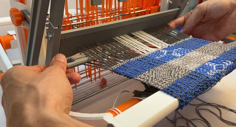Puede incluir: Primer plano de un telar con una tela tejida en tonos de azul, gris y blanco. La tela se est&aacute; tejiendo en un telar con partes naranjas y grises. Se ven las manos de una persona operando el telar.